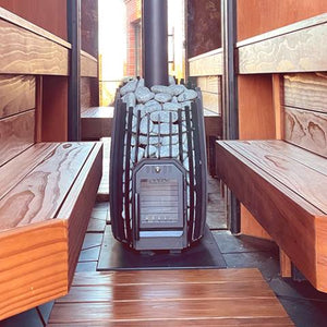 An eye-level, centered view looking down the middle of a wooden sauna room. In the center, a barrel-shaped black metal sauna stove stands filled with light grey stones, mounted on a protective black base plate with a thick black chimney pipe rising straight up from the top. The unlit firebox door faces forward. Flanking the stove on the left and right sides are matching, parallel multi-tiered wooden benches with a prominent natural wood grain.