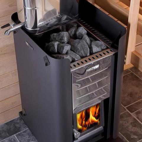 A wood-burning sauna stove enclosed by a matte black protective metal shroud. The stove is filled with dark, rough-textured sauna stones on top. A shiny stainless steel chimney pipe or water tank with a spigot rests on the upper left side. The front of the stove features a polished chrome panel with a "HARVIA" logo and a glass door below revealing bright orange and yellow flames. The setup sits on a grey tiled floor next to light wood paneled walls and a wooden bench.