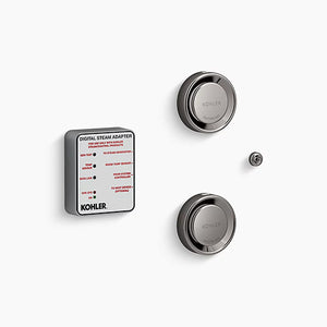 A Kohler digital steam adapter kit on a white background. On the left is a thick, square module with rounded corners, a matte gray body, and a white label face featuring technical text and red indicators. To the right are two titanium, circular steam heads stacked vertically, each featuring a reflective, rounded outer rim and a central dial embossed with "Kohler" and "Caution Hot." A tiny, matching titanium circular sensor button sits on the far right between the two steam heads.