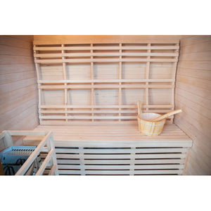 The image shows the interior of the traditional sauna, featuring a slatted wooden bench, a large contoured wooden backrest mounted on the back wall, a wooden bucket and spoon set sitting on the bench, and a protective wooden guard frame around the heater on the lower left.
