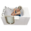 An older woman with grey hair sits inside a white walk-in bathtub, using a handheld shower head to spray water on her shoulder.