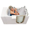An older woman with grey hair sits inside a white walk-in bathtub, using a handheld shower head to spray water on her collarbone and chest.