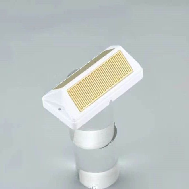 A white rectangular sensor module rests at an angle on a cylindrical stand against a grey background, featuring a prominent gold zigzag grid pattern on its top face.