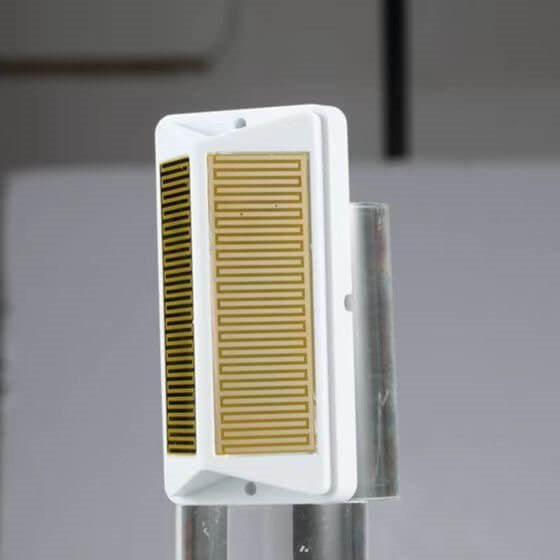 A white rectangular sensor plate stands vertically on a clear glass cylindrical base, showcasing a large gold zigzagging grid pattern and a smaller dark, narrow grid along its left side.