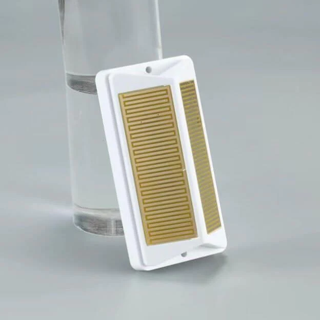 A white rectangular sensor plate leans vertically against a clear glass filled with water on a grey surface, displaying a large gold zigzagging grid pattern and a smaller adjacent grid on its face.