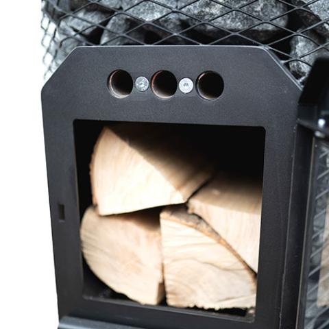 A close-up view shows the open firebox of a Cozy Heat wood-burning sauna stove. The black cast-iron door is swung open to the right side, revealing several large, split pieces of dry firewood neatly stacked inside the dark chamber. Directly above the main opening, the matte black frame features 3 small, circular air vents arranged horizontally, secured w/ two screws. In the background, just above the firebox structure, the wire mesh cage & stacked grey sauna stones are partially visible.