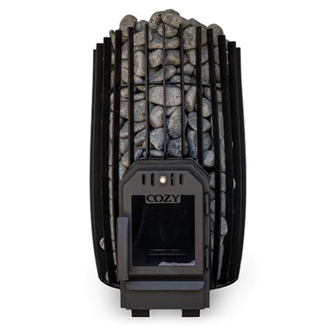 A straight-on view shows a black Cozy Heat sauna stove tightly packed with stones against a white background. The heater features a slightly bowed barrel-style outer cage built from thick vertical black slats. Smooth, rounded grey olivine stones are piled high inside the frame, mounding slightly over the top rim. At the bottom front, a black matte firebox door with a glass viewing window is prominently featured, complete with a vertical handle and the "COZY" brand name stamped above the glass.