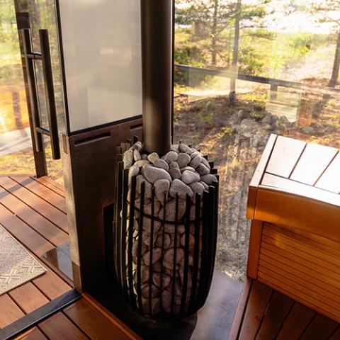 A vertical, barrel-shaped black metal cage stove filled with grey sauna rocks sits on a dark hearth plate. A matte black chimney pipe extends upward from the center of the stones. The stove is positioned next to a glass-paneled wall and door that look out onto a sunny, wooded outdoor landscape. To the right, a warm-toned, smooth wooden sauna bench is visible. Sunlight streams across the rich, slatted wooden floorboards of the sauna interior.