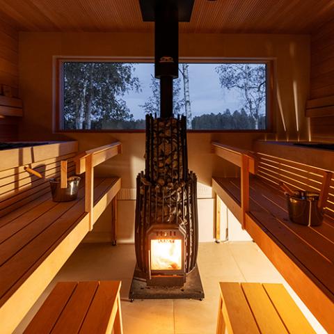 Inside a wooden sauna, a tall black stone-filled heater sits centrally. Its base glows with a bright orange fire visible through a glass door. Multi-tiered, light-colored wooden benches line the left and right walls, holding metal buckets with wooden ladles. In the background, a wide rectangular window looks out onto a serene, dim forest landscape with birch trees under a twilight sky. Warm, indirect yellow light illuminates the clean tile floor from beneath the benches.