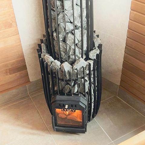 A high-angle view shows a black Cozy Heat sauna stove extension assembly placed on a tiled floor between wood-paneled walls. The unit features a barrel-shaped lower cage made of thick vertical black slats, transitioning into a narrower matching vertical-slat chimney cover cage extending upward. Both sections are tightly packed with jagged, grey split-face stones. At the bottom front, a black metal firebox door with a glass window reveals lit wood burning inside.