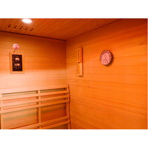 The interior of the sauna features warm wooden wall paneling, a digital control panel mounted on the left wall above a slatted backrest, a vertical thermometer strip, and a circular analog hygrometer/thermometer gauge mounted on the right wall.