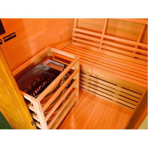 The image shows an interior view of the sauna featuring a Harvia heater enclosed within a protective wooden guard frame, positioned next to a wooden bench with a slatted backrest and warm wood flooring.