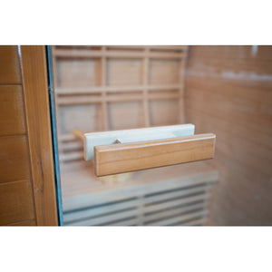 The image shows a close-up view of the wooden door handle mounted on the glass door of the sauna, looking through into the interior featuring a slatted bench and backrest.