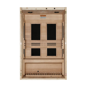 A straight-on view looking directly into the open front of a light-colored wood infrared sauna cabin. The interior back wall features four large, vertical rectangular black heating panels arranged in pairs (two upper, two lower), partially divided by horizontal wooden backrest slats. A wide wooden bench spans the width of the lower section, below which is a matching light wood slatted floor mat. The top ceiling panel has small circular cutouts, and a warning label is affixed above the panels.