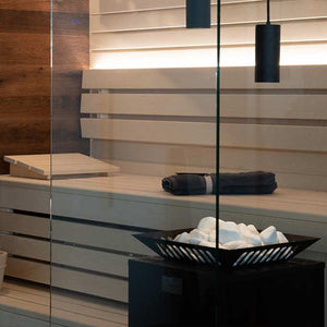 Sauna interior with wood slat benches, backlit wall panel, rolled towel, wooden headrest, glass door, and black heater with stone tray in foreground.