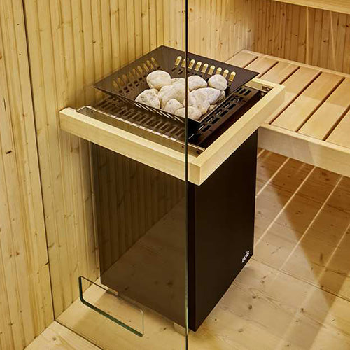 Black EOS sauna heater with wood-trimmed top and stone tray filled with white sauna rocks, installed in wood-paneled sauna with slatted bench and glass door.