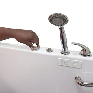 Close-up of a hand adjusting a control dial on the rim of a white walk-in bathtub, with chrome handheld shower, faucet handle, control panel, and grab bar visible.