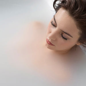 Woman with eyes closed relaxing in a bath, close-up of her face and shoulders submerged in milky-white water.