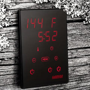 A Harvia digital sauna control panel displays "144 F" and "5:52" in red LED text. The black touch panel is mounted on a rustic wood surface and features illuminated red icons for temperature, time, power, and menu controls.
