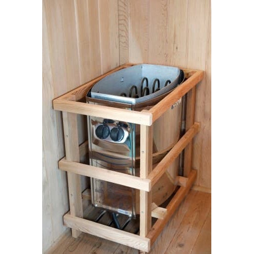 A close-up shot of a Harvia traditional sauna heater unit installed in the corner of an interior sauna cabin, featuring a metallic finish, control dials, exposed heating elements, and a protective wooden safety fence enclosure.