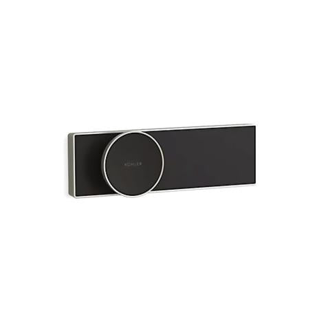 A sleek, modern Kohler control panel on a solid white background. The unit consists of a long, thin rectangular plate with a matte black front face and a brushed nickel metallic border. Positioned off-center to the left is a large, circular control dial that slightly overlaps the top and bottom edges of the rectangle. The dial has a matching matte black face, a thin brushed nickel rim, and the word "KOHLER" written in small, crisp white capital letters across its center.