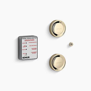 A Kohler digital steam adapter kit on a white background. On the left is a thick, square module with rounded corners, a matte gray body, and a white label face featuring technical text and red indicators. To the right are two gold, circular steam heads stacked vertically, each featuring a reflective, rounded outer rim and a central dial embossed with "Kohler" and "Caution Hot." A tiny, matching gold circular sensor button sits on the far right between the two steam heads.