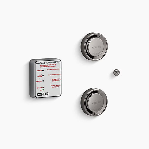A Kohler digital steam adapter kit on a white background. On the left is a thick, square module with rounded corners, a matte gray body, and a white label face featuring technical text and red indicators. To the right are two titanium, circular steam heads stacked vertically, each featuring a reflective, rounded outer rim and a central dial embossed with "Kohler" and "Caution Hot." A tiny, matching titanium circular sensor button sits on the far right between the two steam heads.