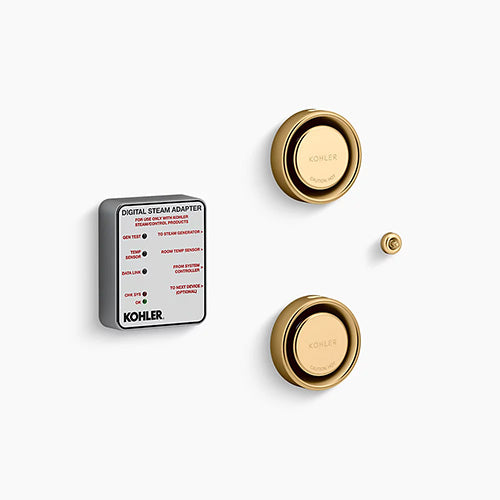 A Kohler digital steam adapter kit on a white background. On the left is a thick, square module with rounded corners, a matte gray body, and a white label face featuring technical text and red indicators. To the right are two brushed brass, circular steam heads stacked vertically, each featuring a reflective, rounded outer rim and a central dial embossed with "Kohler" and "Caution Hot." A tiny, matching brushed brass circular sensor button sits on the far right between the two steam heads.