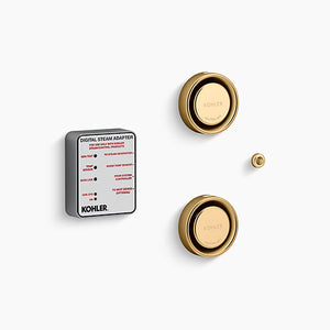 A Kohler digital steam adapter kit on a white background. On the left is a thick, square module with rounded corners, a matte gray body, and a white label face featuring technical text and red indicators. To the right are two brushed brass, circular steam heads stacked vertically, each featuring a reflective, rounded outer rim and a central dial embossed with "Kohler" and "Caution Hot." A tiny, matching brushed brass circular sensor button sits on the far right between the two steam heads.