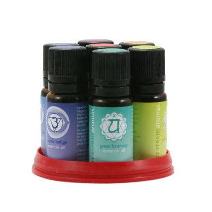 A product photo of the Mr. Steam Multi Chakra Aroma Oil Package against a white background, featuring a line-up of seven 10 mL amber glass bottles with black caps and solid colored labels (red, orange, green, turquoise, blue, dark blue, and purple) displaying chakra symbols, accompanied by a fan of matching colored circular labels or cards arranged in the foreground.