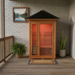 The SunRay Bristow 2-Person Outdoor Wet/Dry Traditional Sauna (HL200D2) is positioned on a covered wooden porch against a white brick wall. The setup features the compact wooden sauna with its pyramid-style roof, flanked by potted plants—a lush fern on the left and a tall snake plant on the right. A welcoming doormat sits right in front of the glass entry door, and a framed nature landscape picture hangs on the brick wall above the left side of the sauna.
