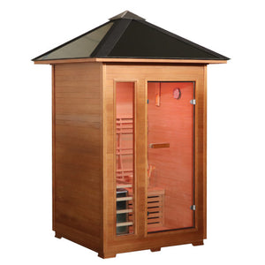 The SunRay Bristow 2-Person Outdoor Wet/Dry Traditional Sauna with 4.5kW Harvia Heater (HL200D2) features a compact wooden construction with a distinct pyramid-style pitched metal roof and overhang. The front facade includes a glass entry door next to a vertical side window, providing a clear view of the interior wooden benches, slatted backrests, and lighting fixtures.