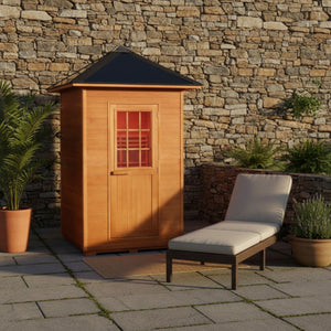 The SunRay Eagle 2-Person Outdoor Traditional Wet/Dry Sauna (HL200D1) is set up on a stone patio against a rustic dry-stone wall. The outdoor relaxation area includes the wooden sauna featuring its pyramid-style roof and multi-pane window door, complemented by a padded wicker chaise lounge chair, a welcoming doormat, and potted greenery on both sides.