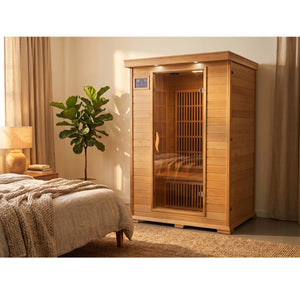 The SunRay Evansport 2-Person Indoor Infrared Sauna (model HL200K2) installed in a cozy, warmly lit bedroom with a wooden floor, a bed with a knit blanket in the foreground, a potted fiddle-leaf fig tree next to the sauna, and sheer curtains framing a window.