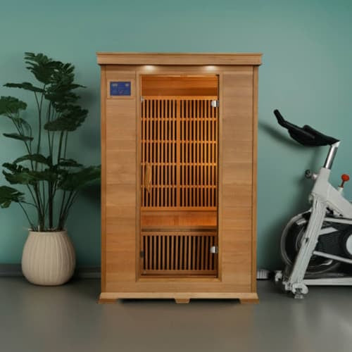 An indoor light cedar wood infrared sauna stands centrally against a solid teal-green wall on a gray floor. The sauna features a front glass door with a digital control panel on the upper left and slatted wooden interior backrests. To the left is a tall potted plant in a round white pot, and to the right is an exercise stationary bike.