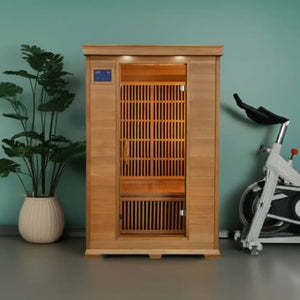 An indoor light cedar wood infrared sauna stands centrally against a solid teal-green wall on a gray floor. The sauna features a front glass door with a digital control panel on the upper left and slatted wooden interior backrests. To the left is a tall potted plant in a round white pot, and to the right is an exercise stationary bike.
