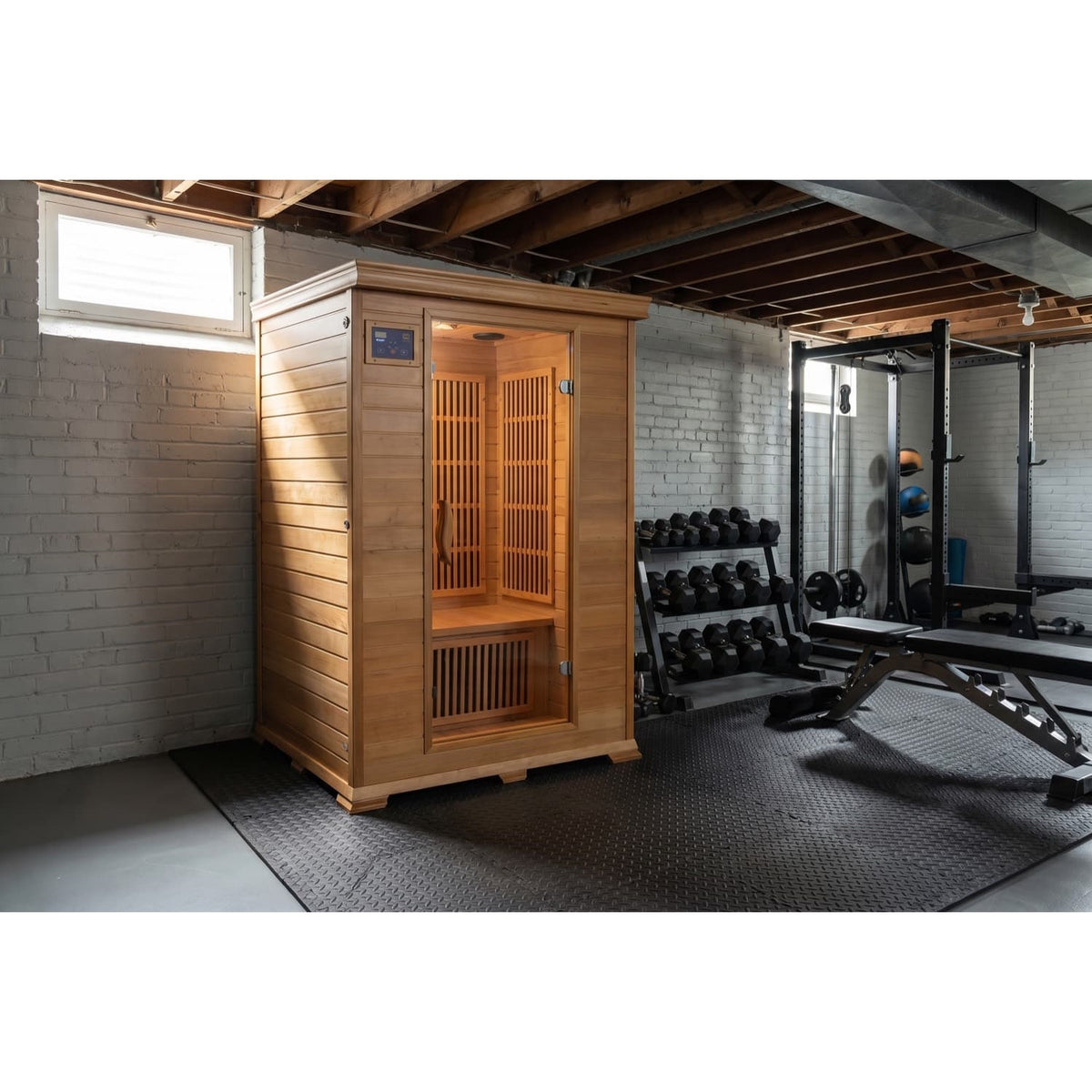 The SunRay Evansport 2-Person Indoor Infrared Sauna (model HL200K2_6) set up in a home gym basement with white painted brick walls, exposed ceiling joists, a concrete floor, and fitness equipment including a dumbbell rack and weight bench nearby.
