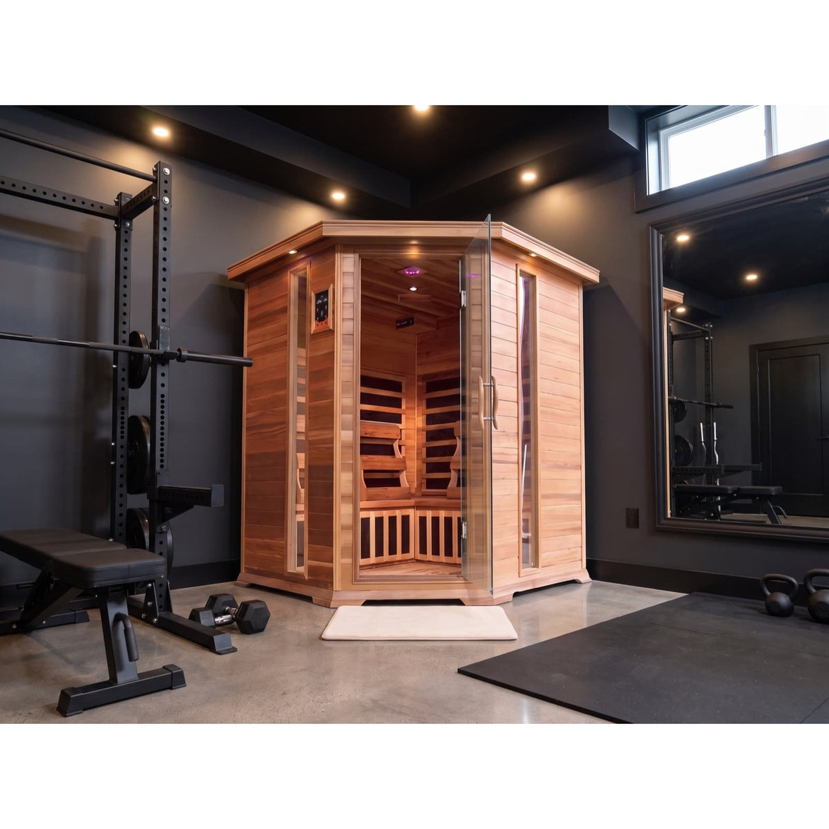 The SunRay Sauna Bristol Bay Indoor FAR Infrared Corner Sauna (model HL400KC_1) installed in a modern home gym with dark charcoal walls, concrete flooring, a weight rack, and dumbbells.