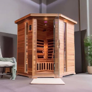 The SunRay Sauna Bristol Bay Indoor FAR Infrared Corner Sauna (model HL400KC_2) installed in a room with smooth gray flooring and walls, featuring a hexagonal wooden corner design, horizontal cedar planks, a glass door with side windows, and a chair with a draped blanket nearby.