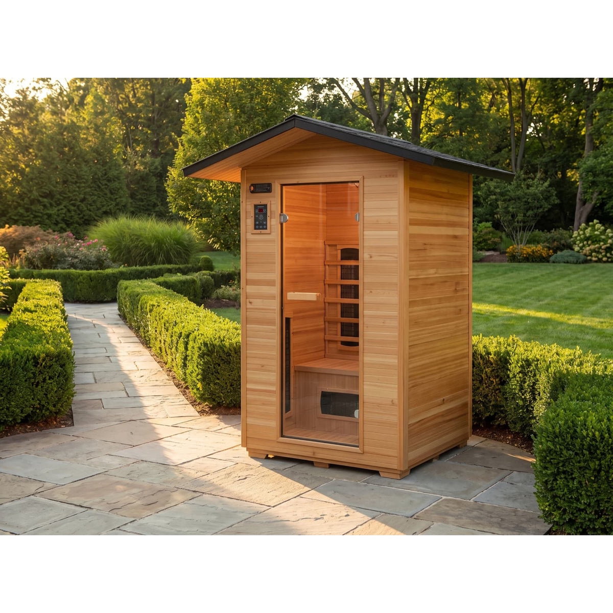The SunRay Sauna Burlington 2-Person Outdoor Infrared Sauna (model HL200D3) installed on a stone patio in a lush backyard garden setting, featuring a weather-resistant wooden cabin structure, an overhanging shingled roof, a glass door, and exterior digital controls.