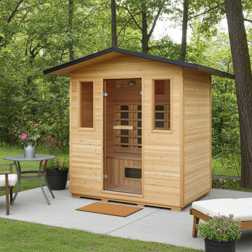 The SunRay Sauna Cayenne 4-Person Outdoor Infrared Sauna (HL400D3) is nestled in a serene wooded backyard setting on a paved patio. The wooden outdoor sauna features a pitched metal roof with an overhang, a glass entry door flanked by two vertical windows, and a welcome mat at the entrance. A small patio table with flowers and an outdoor lounger complete the tranquil relaxation area.
