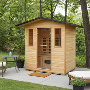 The SunRay Sauna Cayenne 4-Person Outdoor Infrared Sauna (HL400D3) is nestled in a serene wooded backyard setting on a paved patio. The wooden outdoor sauna features a pitched metal roof with an overhang, a glass entry door flanked by two vertical windows, and a welcome mat at the entrance. A small patio table with flowers and an outdoor lounger complete the tranquil relaxation area.