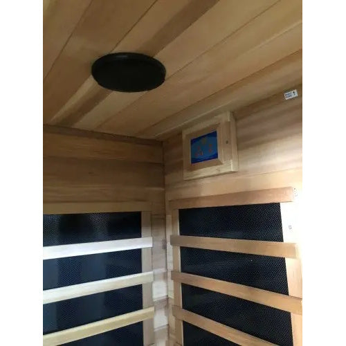 An interior perspective of the SunRay Savannah 3-Person Infrared Cedar Sauna (model HL300K_3), showing a dark round speaker mounted on the cedar wood ceiling and adjacent interior walls featuring infrared heating panels behind slatted guards and a control panel.