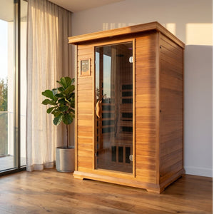 The SunRay Sierra 2-Person Indoor Infrared Cedar Sauna (model HL200K) installed in a bright room with a wooden floor, next to a large window with sheer curtains and a potted fiddle-leaf fig tree.