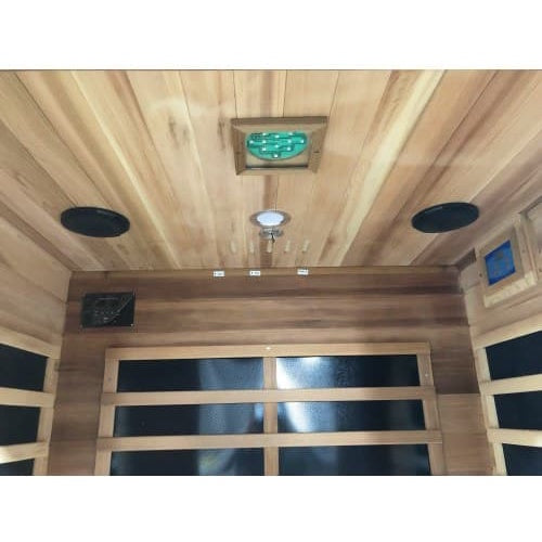 An interior perspective of the SunRay Sierra 2-Person Indoor Infrared Cedar Sauna (model HL200K_4), showing the cedar wood ceiling fitted with two black round speakers, a central light fixture, and a square chromatherapy lighting panel, looking down toward the back wall with infrared heating panels.