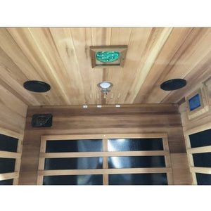 An interior perspective of the SunRay Sierra 2-Person Indoor Infrared Cedar Sauna (model HL200K_4), showing the cedar wood ceiling fitted with two black round speakers, a central light fixture, and a square chromatherapy lighting panel, looking down toward the back wall with infrared heating panels.