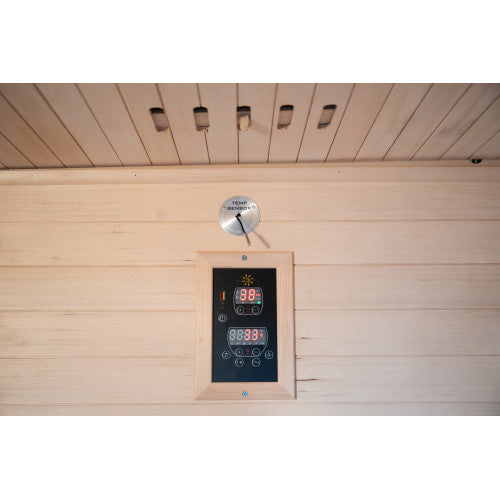The image shows components mounted on the interior wooden wall and ceiling paneling of the sauna, featuring a digital control panel framed in wood, a labeled metal temperature sensor plate positioned just above it, and a row of ventilation slots along the ceiling panel.
