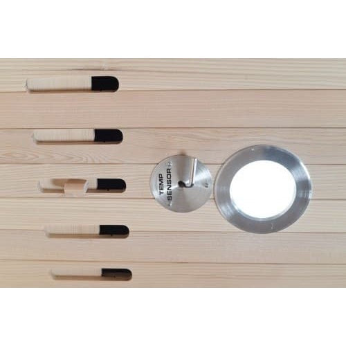 A close-up shot of an interior sauna wall made of light wood planks, featuring a circular silver-metallic temperature sensor plate with a hook, a round embedded light fixture with a metal bezel, and a series of horizontal ventilation slots with adjustable wooden covers on the left.