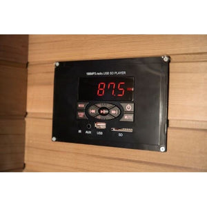 A close-up shot of a black digital USB/MP3/radio media player console flush-mounted on a horizontal wooden sauna wall, featuring an LED display showing a radio frequency, control buttons, and USB, SD card, and AUX ports.