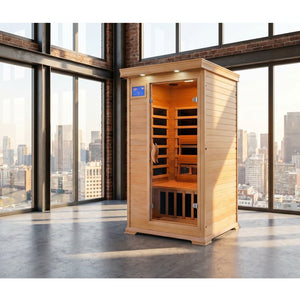 An indoor, light-brown cedar infrared sauna stands in the middle of a spacious, modern loft with a polished concrete floor and large floor-to-ceiling windows offering a city skyline view. The sauna features a glass door, a digital control panel on the upper left, interior bench seating, and dark heating panels.